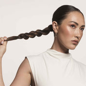 A woman with smooth, dark hair in a twisted ponytail—styled with No Frizz Smooth Styling Spray—wears a sleeveless white top and small hoop earrings. She gazes to the side, holding her braided ponytail against a light background.
