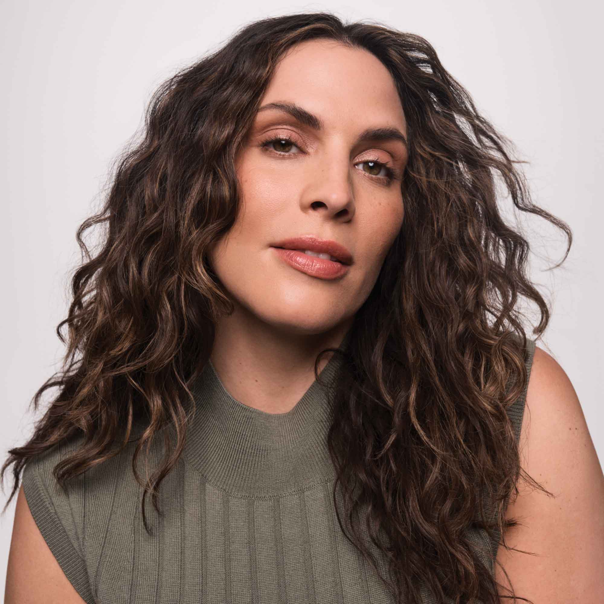 A woman with long, wavy brown hair and natural texture, wearing a sleeveless olive-green top, showcases her confidence and effortless style—her look enhanced by Full Texturizing Foam—against a neutral light background.