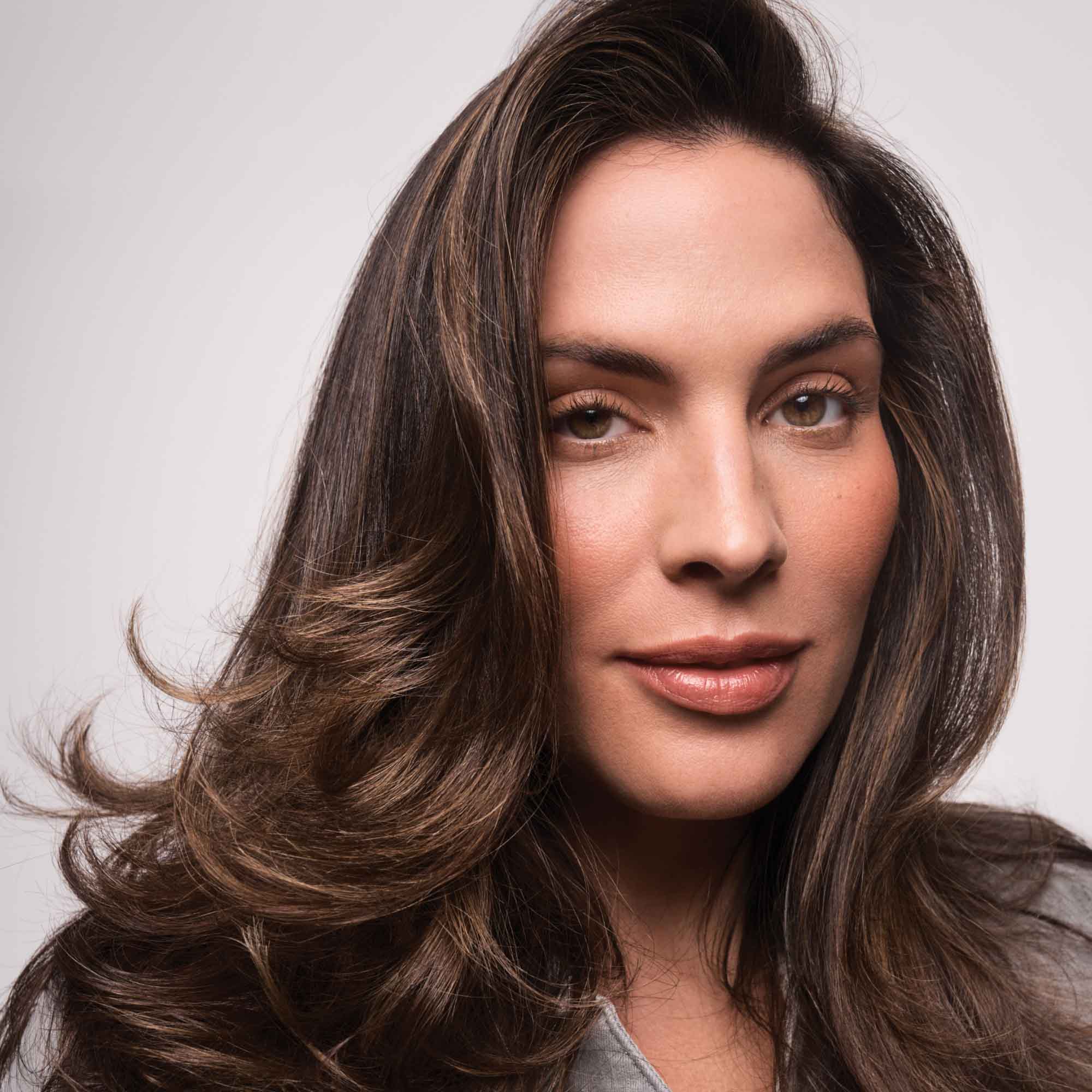 A woman with long, wavy brown hair and natural makeup looks confidently at the camera against a plain light background, her style elevated by Full Volume & Root-Lifting Spray for all-day fullness.