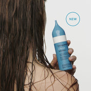 A person with wet, brown hair holds a blue and white bottle of Scalp Care Exfoliator. The pointed applicator tip targets dry scalp flaking. A blue circle labeled NEW is in the top right corner.