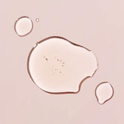 Three clear drops of Curl Moisturizing Shine Oil, inspired by biomimetic oil that mimics hair’s natural lipids, are arranged on a light pink surface; the largest drop contains small bubbles inside.