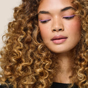 With voluminous, curly light brown hair and glowing skin, they look downward. Wearing light purple eyeliner, pink blush, and natural pink lipstick, their curls shine with Curl Moisturizing Shine Oil against a neutral background.