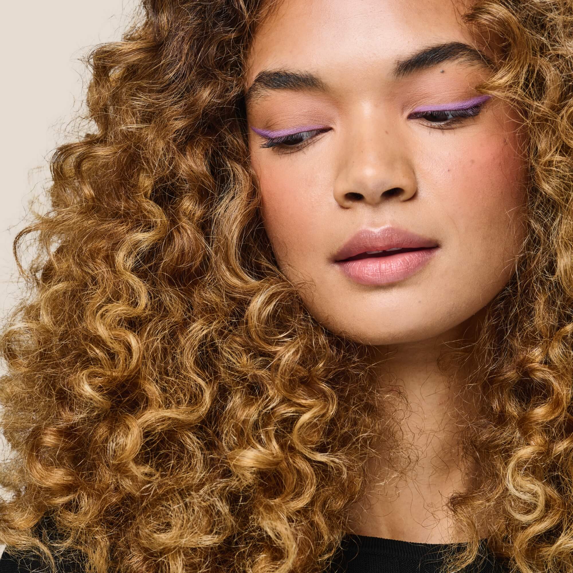 With voluminous, curly light brown hair and glowing skin, they look downward. Wearing light purple eyeliner, pink blush, and natural pink lipstick, their curls shine with Curl Moisturizing Shine Oil against a neutral background.