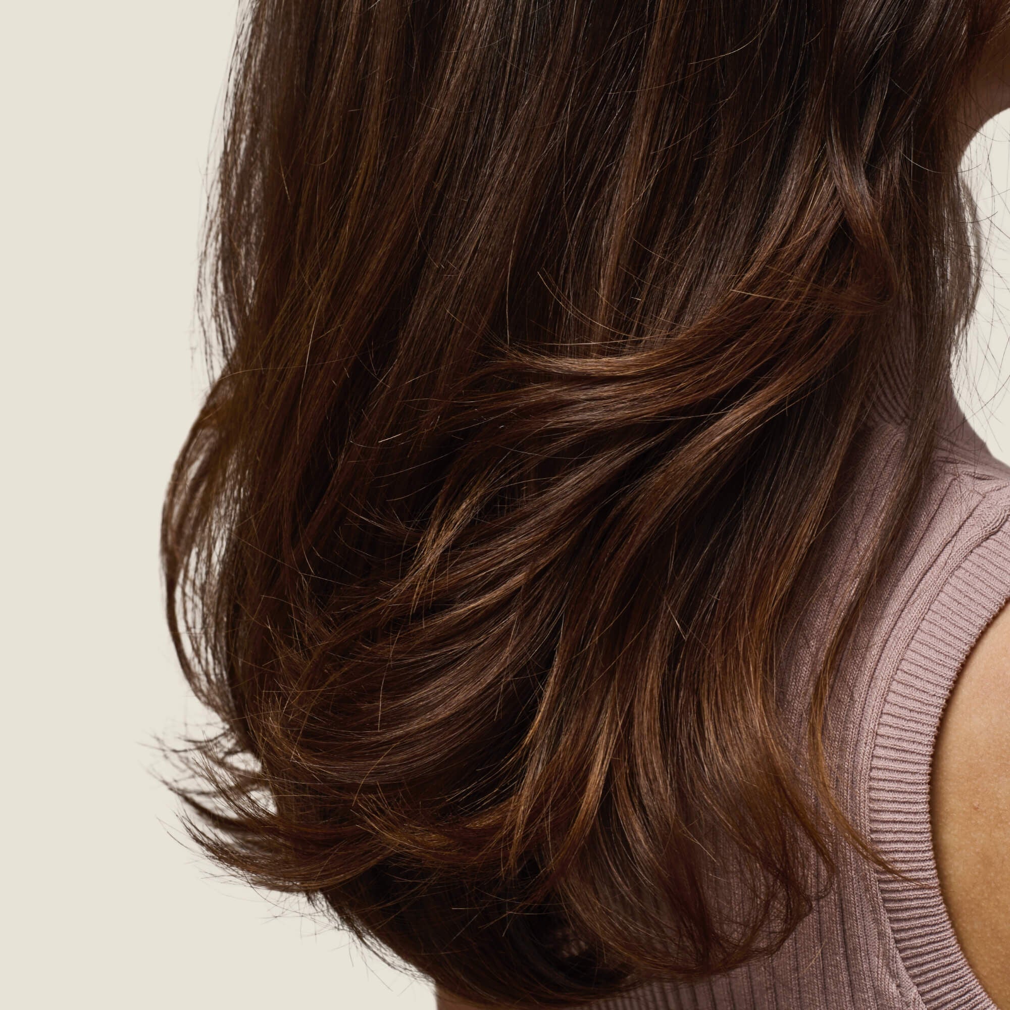 A woman with long, smooth, naturally voluminous dark brown hair—achieved with Full Thickening Mousse—wears a sleeveless ribbed mauve top and is shown from the back and side against a plain light background.