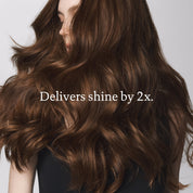 A woman with long, shiny, wavy brown hair wears a black top and faces slightly away. Text over the image reads: Curl Curly Hair Essentials delivers 2x added shine and frizz-free curls.