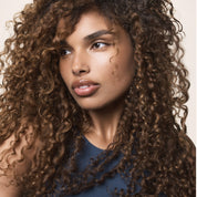 A woman with long, voluminous, curly brown hair—freshly washed with Clarifying Detox Shampoo—looks to the side. Her smooth skin, defined brows, and glossy lips complement her sleeveless dark blue top against a light background.