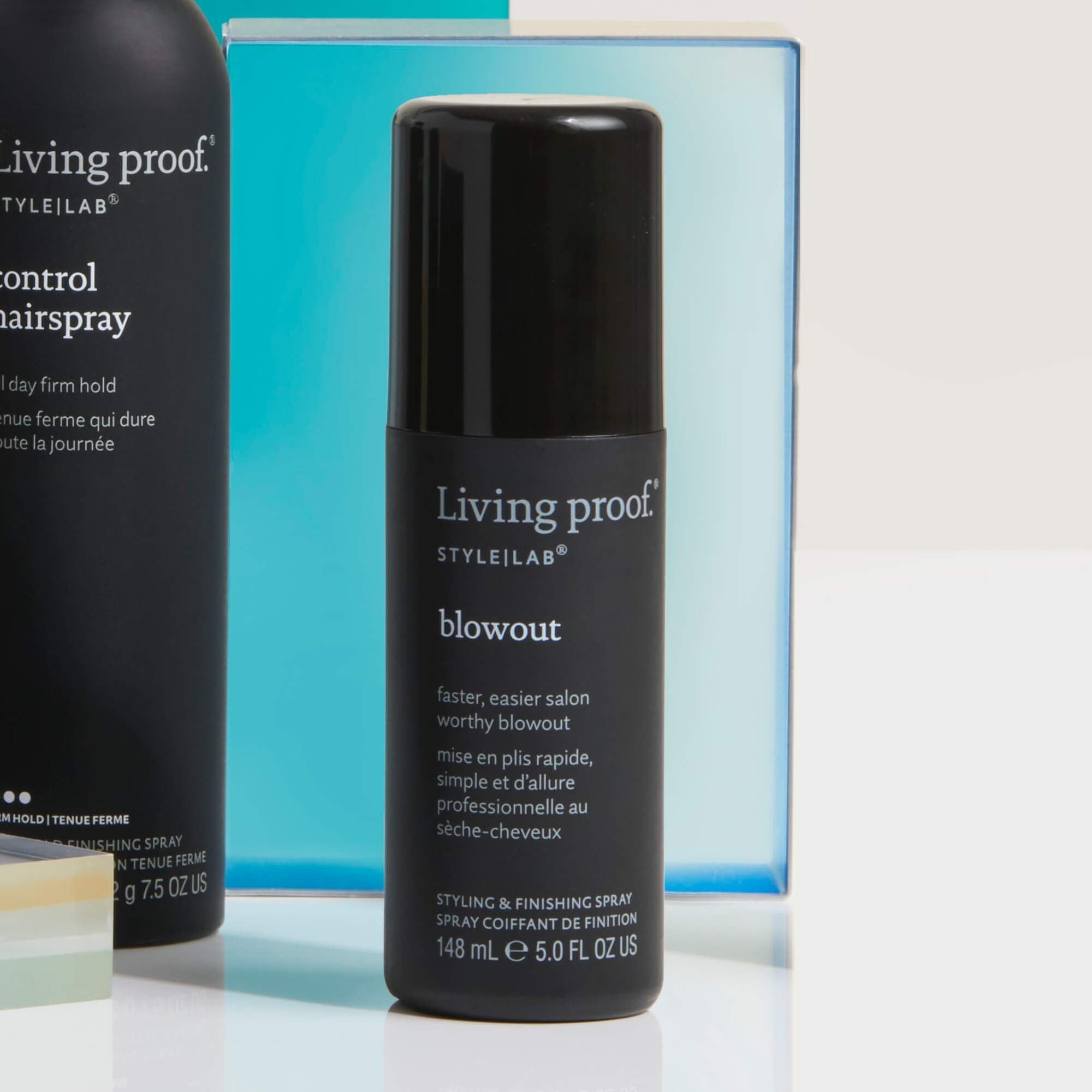 A black bottle of Style Lab® Blowout styling spray stands upright, with a blue box and another Living Proof item partially visible in the background—perfect for achieving a long-lasting blowout.