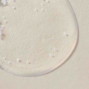 A close-up of a clear, glossy No Frizz Smooth Styling Serum droplet with small bubbles on a textured, light beige surface.