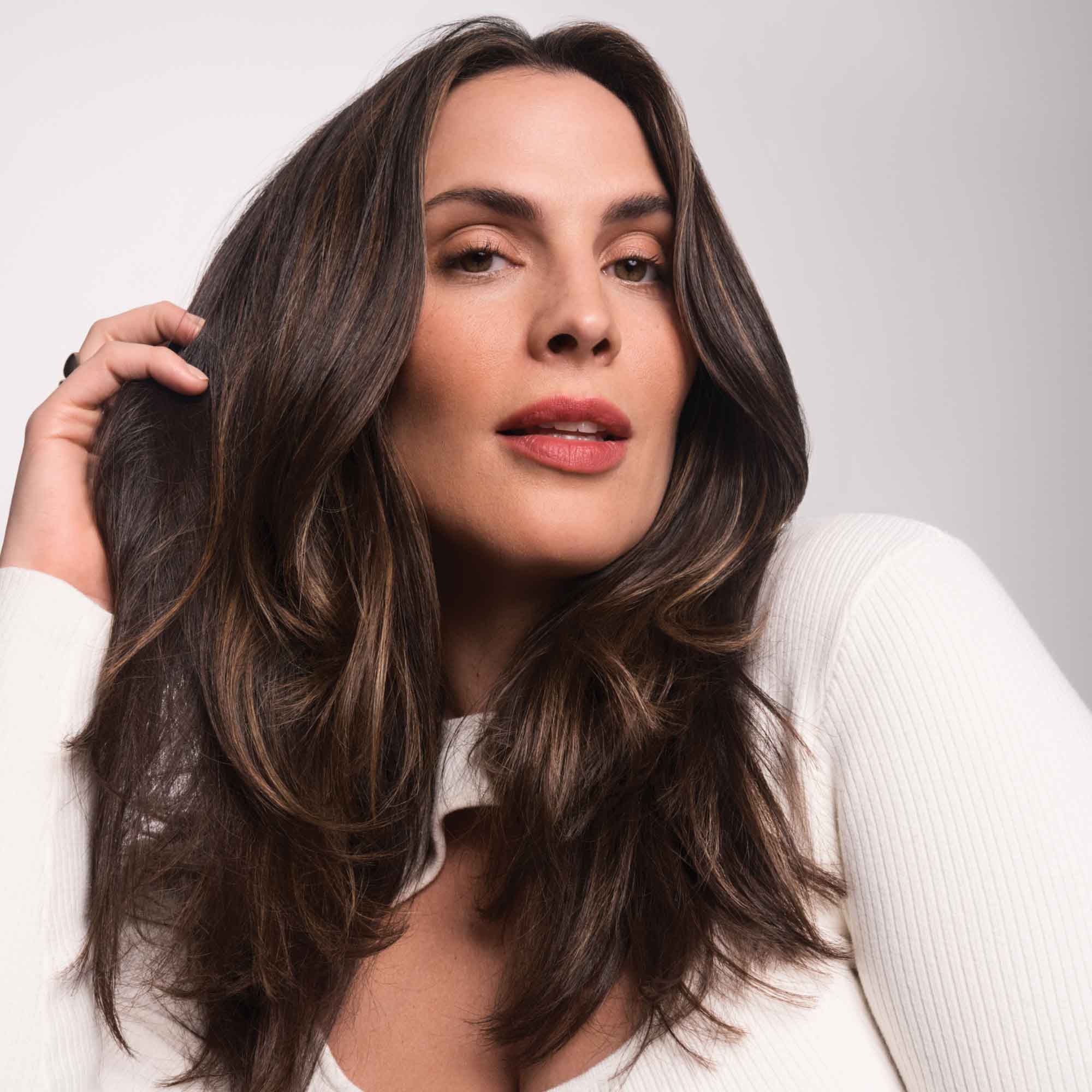 A woman with long, wavy brown hair and natural makeup poses in a white top against a plain background, touching her hair to highlight its fuller look achieved with Full Thickening Blow-Dry Cream.