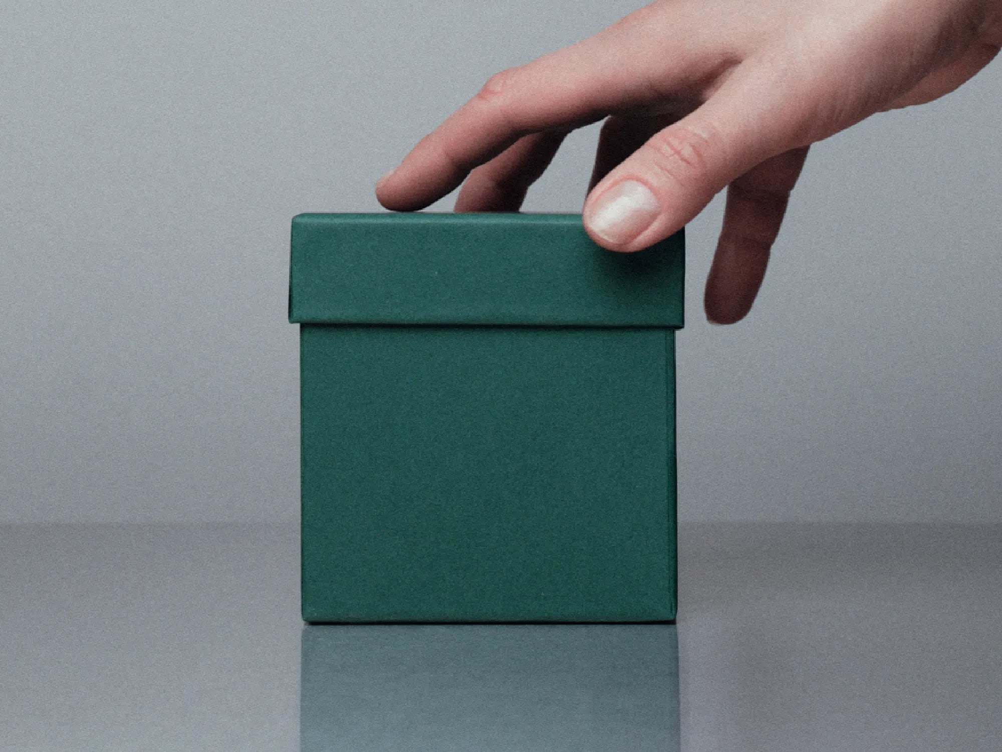Hand reaching towards a green gift box on a gray surface with a neutral background