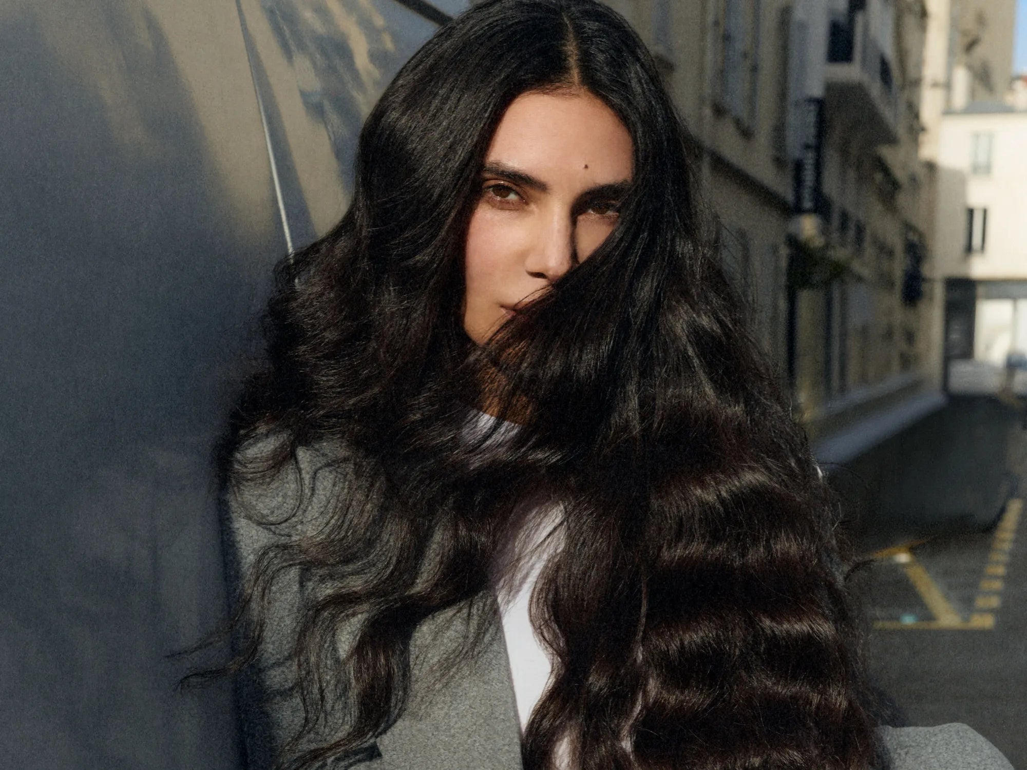 A woman with very long, dark, wavy hair wearing a gray blazer over a white top, photographed outdoors against a blurred urban street background.