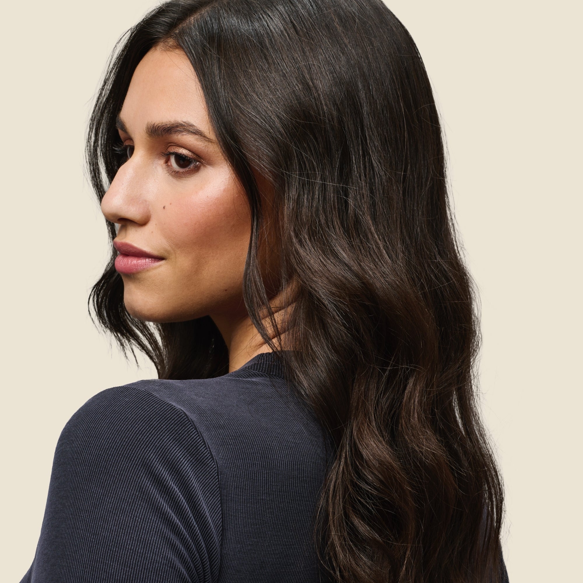 A woman with long, shiny dark brown hair and light skin looks over her shoulder against a beige background, showcasing the effects of No Frizz Vanishing Oil.