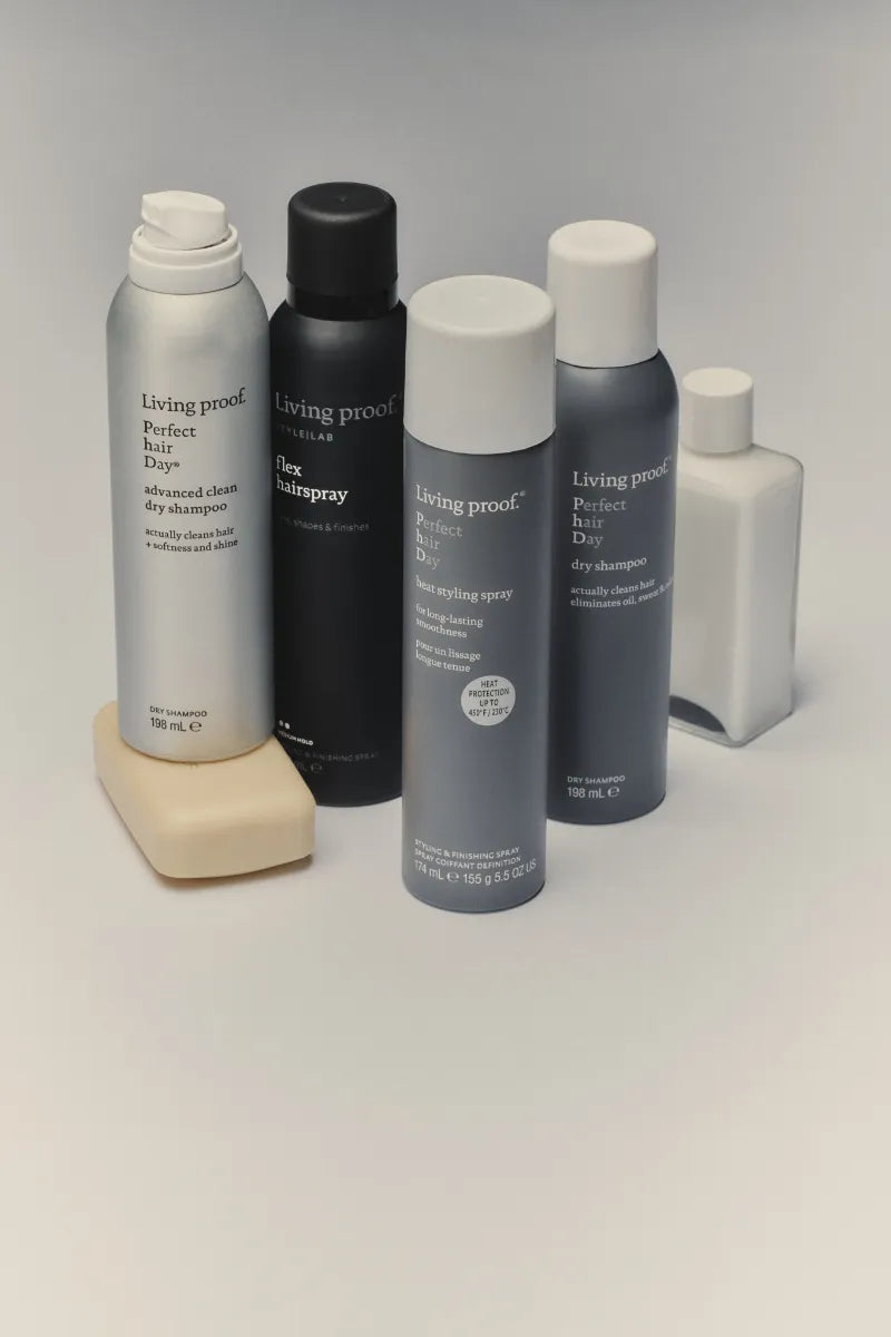 Five Living Proof haircare bottles arranged on a light gray studio background, including a silver shampoo bottle on a beige stand, a black hairspray bottle, a gray hair styling spray bottle, a dark gray dry shampoo bottle, and a small white bottle.