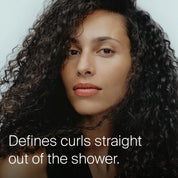 A person with long, defined curly hair and a neutral expression looks at the camera. Text on the image reads: Curl Conditioner hydrates and defines curls straight out of the shower.