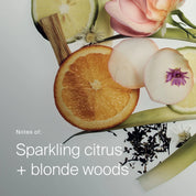 Slices of orange and lime, delicate petals, and wood are arranged on a pale backdrop. Text reads: Curl Conditioner—notes of sparkling citrus and blonde woods, inspiring a hydrating formula that helps define your curls.