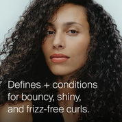 A person with long, dark, shiny curly hair looks at the camera with a neutral expression. Text on the image reads: Curl Definer shapes curls for bounce, shine, and frizz-free definition.