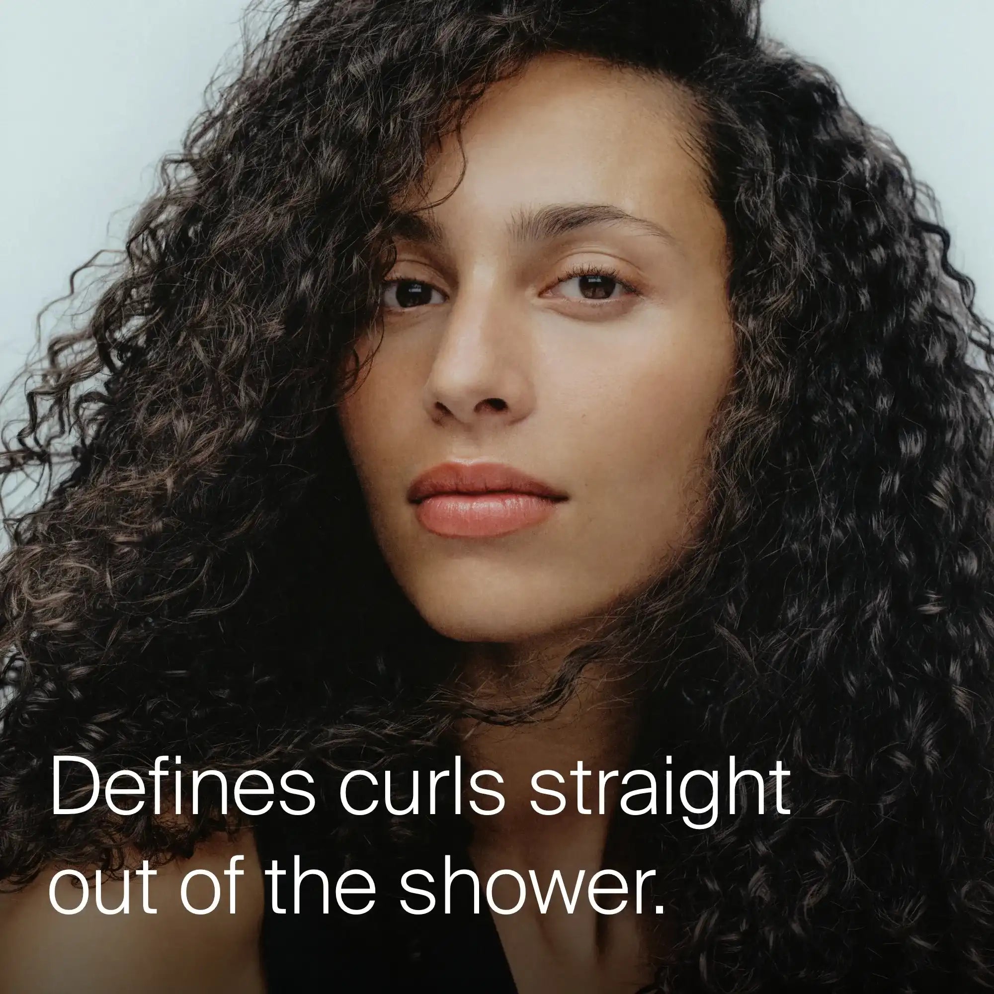 A person with long, defined, dark curly hair looks at the camera with a neutral expression. Text reads: Curl Shampoo—silicone-free, luxe formula defines curls right out of the shower with low lather.