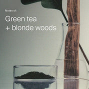 Glass with green tea leaves and wooden spoon on a light gray background with text 'Notes of: Green tea + blonde woods'.
