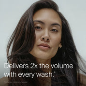A woman with long, voluminous, dark hair looks into the camera. Text reads: Full Conditioner delivers 2x the volume with every wash.* Featuring Sili-CLONE HairTech to improve texture. The background is light and simple.