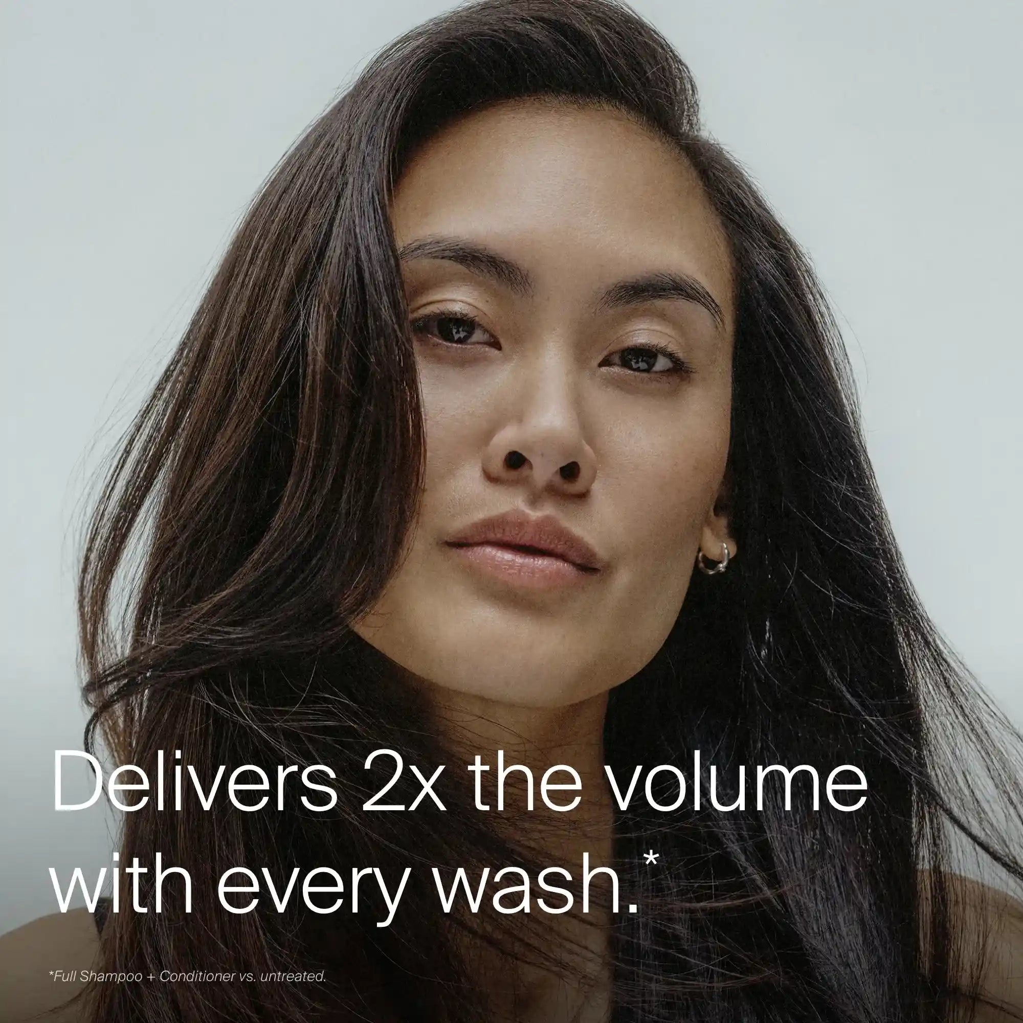 A woman with long, voluminous, dark hair looks into the camera. Text reads: Full Conditioner delivers 2x the volume with every wash.* Featuring Sili-CLONE HairTech to improve texture. The background is light and simple.