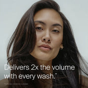 A woman with long, voluminous dark hair looks at the camera. Text reads: Full Shampoo delivers 2x the volume with every wash.* Powered by Sili-CLONE HairTech for noticeable improvement. Neutral, minimal background.