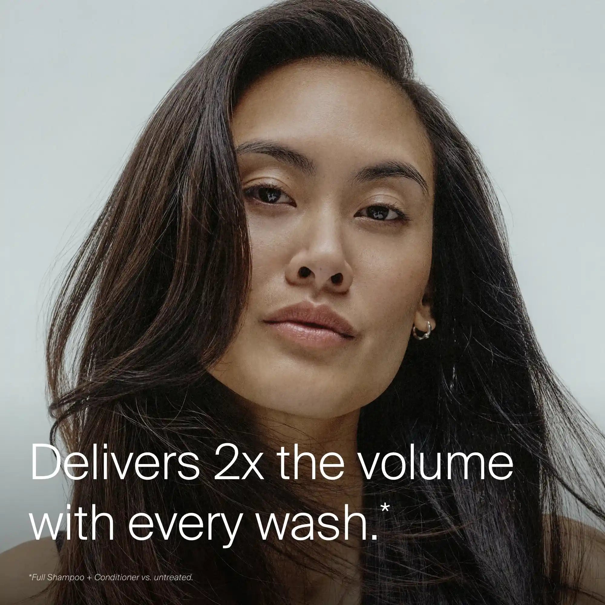 A woman with long, voluminous dark hair looks at the camera. Text reads: Full Shampoo delivers 2x the volume with every wash.* Powered by Sili-CLONE HairTech for noticeable improvement. Neutral, minimal background.