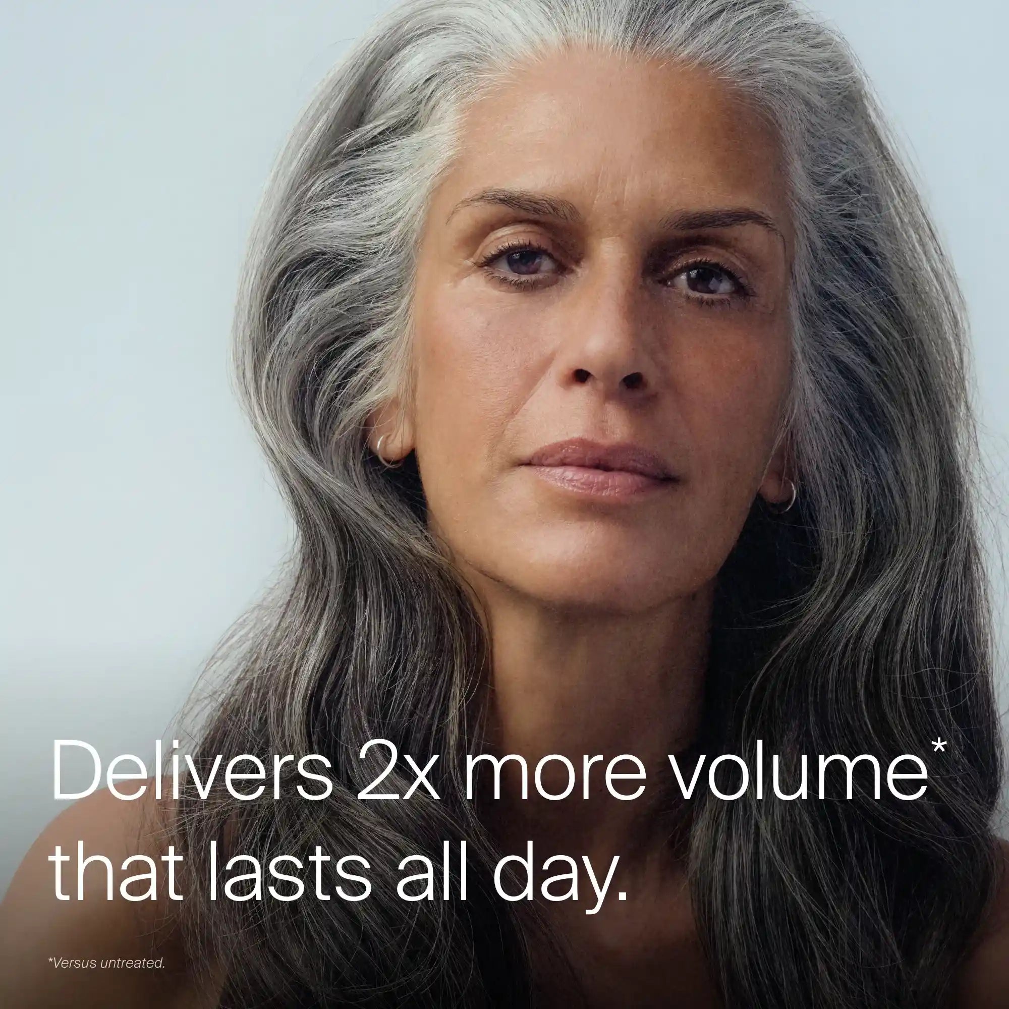 A woman with long, thick, gray hair and a calm expression looks at the camera. Text on the image reads: Full Volume & Root-Lifting Spray delivers 2x more all-day volume* with invisible volume spray. *Vs. untreated hair.