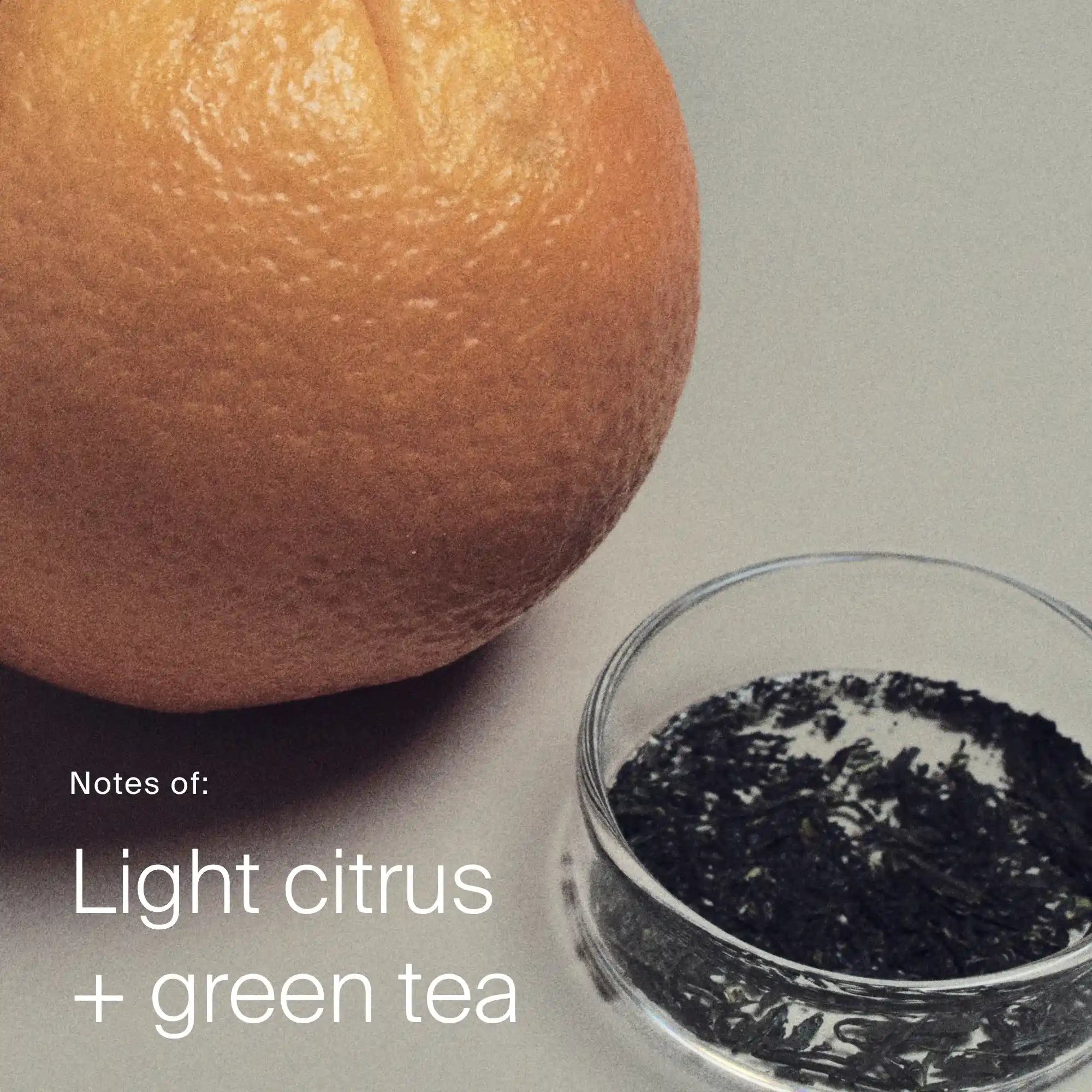 Orange and small bowl of green tea leaves on a neutral background with text 'Notes of: Light citrus + green tea'.