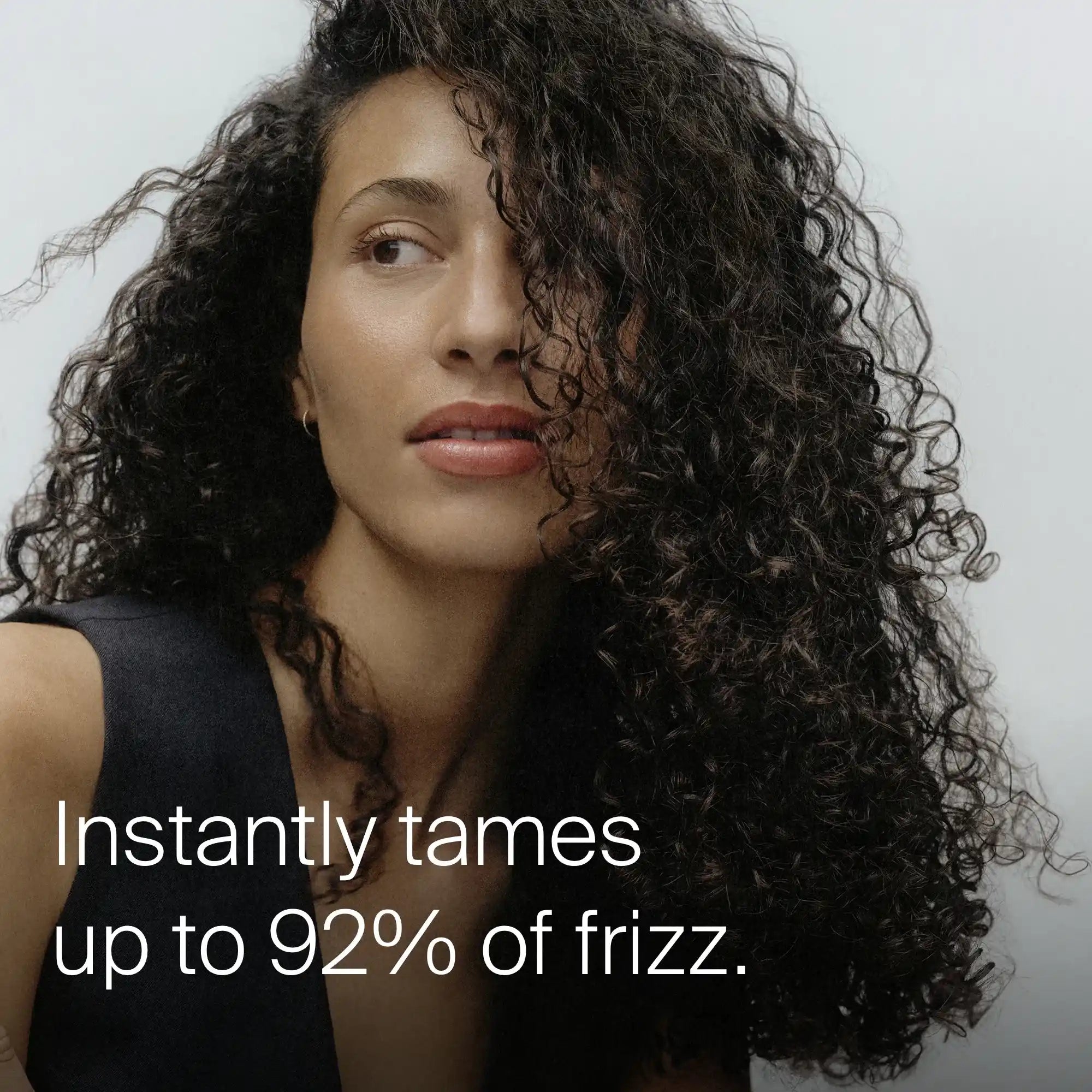 A woman with voluminous, curly hair looks to the side and smiles slightly; text on the image reads: No Frizz Instant De-Frizzer instantly tames up to 92% of frizz for lasting control and irresistible shine.