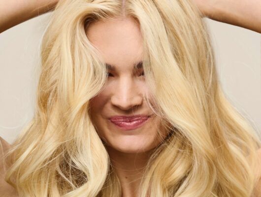 Up close image of a smiling woman with wavy blond hair in front of a beige background.