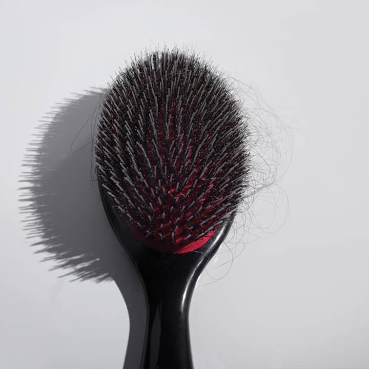 Black and red bristle brush with strands of dark hair in the bristles in front of a gray background.
