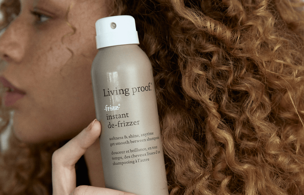 A woman with curly brown hair holding a bottle of Living Proof instant de-frizzer