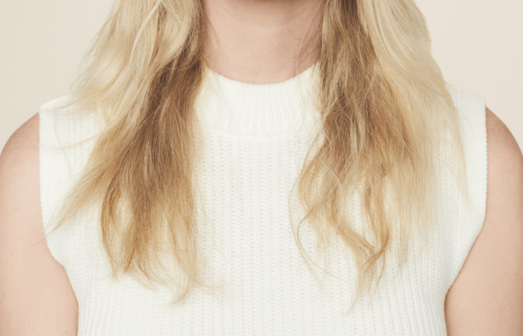 a blonde girl in a white knitted sweater vest photographed from the chin down showing her dry hair.