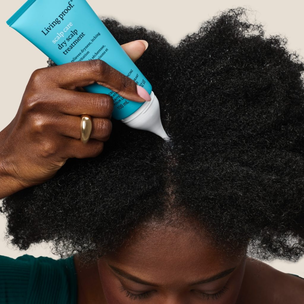 A close-up view of a woman with thick, curly hair applying Living Proof dry scalp treatment to her scalp.