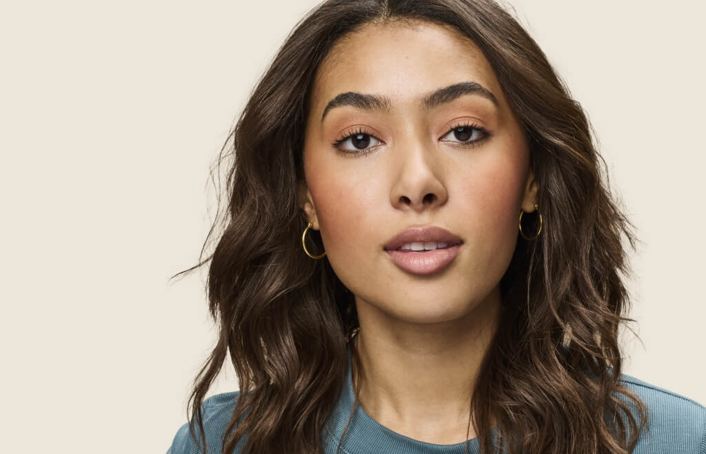 a woman with wavy shoulder length brown hair and gold hoop earrings in front of a beige background