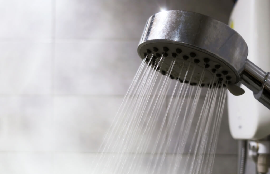 A close-up view of a shower head with water spraying downward in a grey tiled shower filled with steam.
