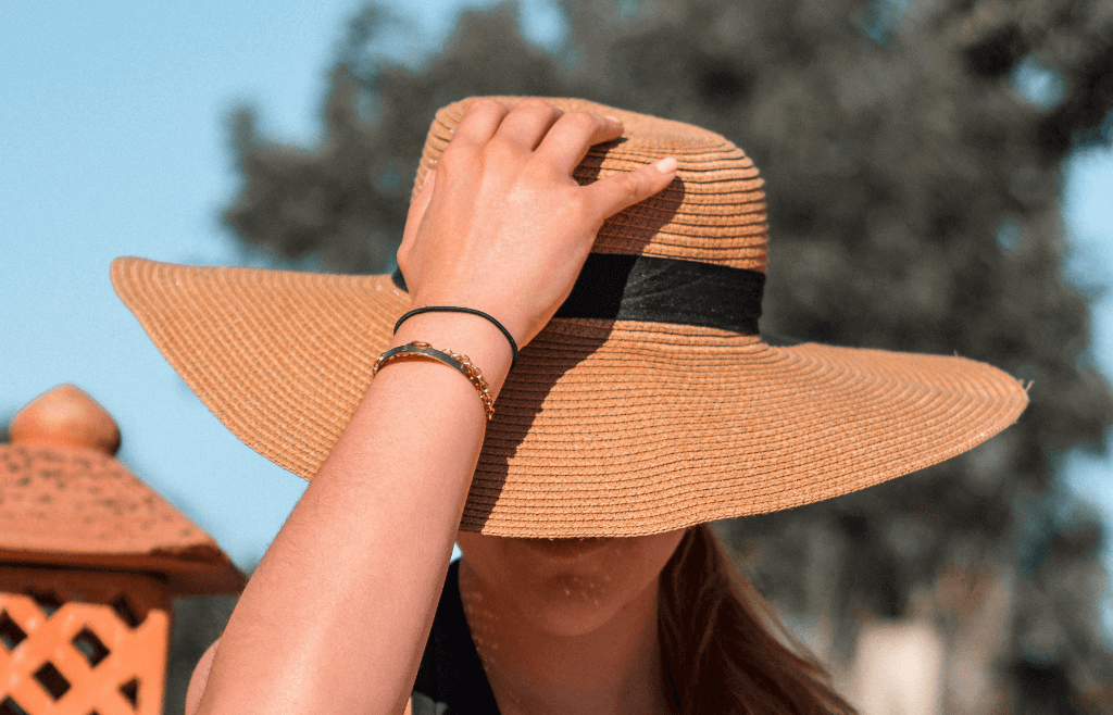 A woman with a tan sun hat on. With her hand on top of the hat looking downwards.