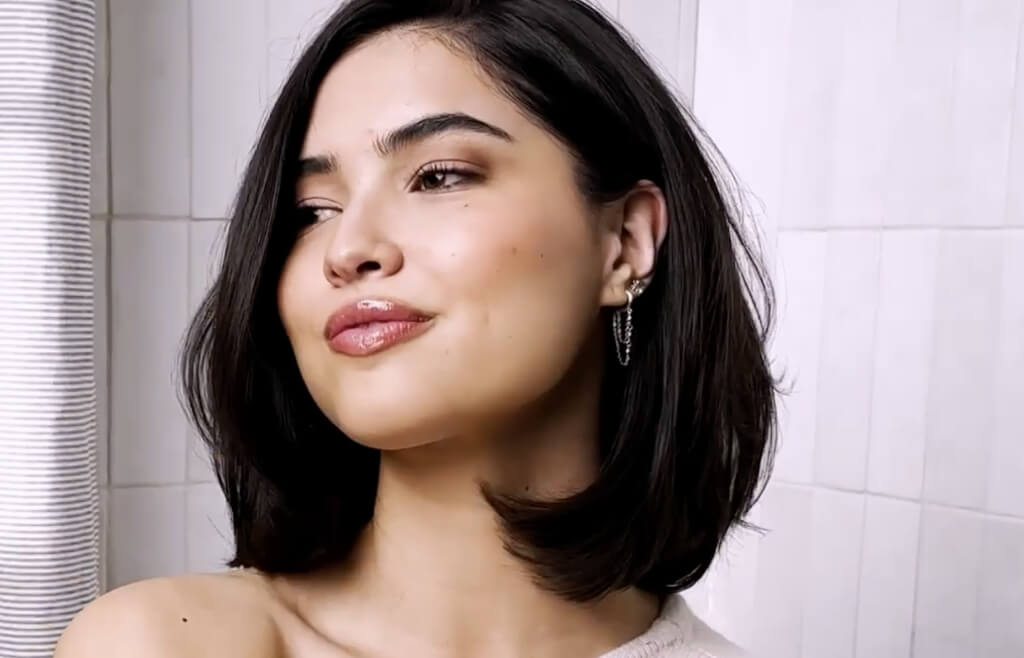 a woman with a black hair cut into a lob stands in front of a white tile wall