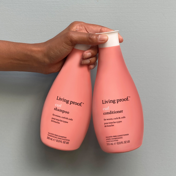 Close-up view of a person holding two coral-colored Living Proof product bottles that hydrate curly hair: Curl Shampoo and Curl Conditioner.